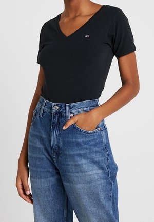 Frau trägt ein schwarzes V-Ausschnitt-Kurzarmshirt, das in eine hoch taillierte blaue Jeans gesteckt ist, mit einer Hand in der Tasche.