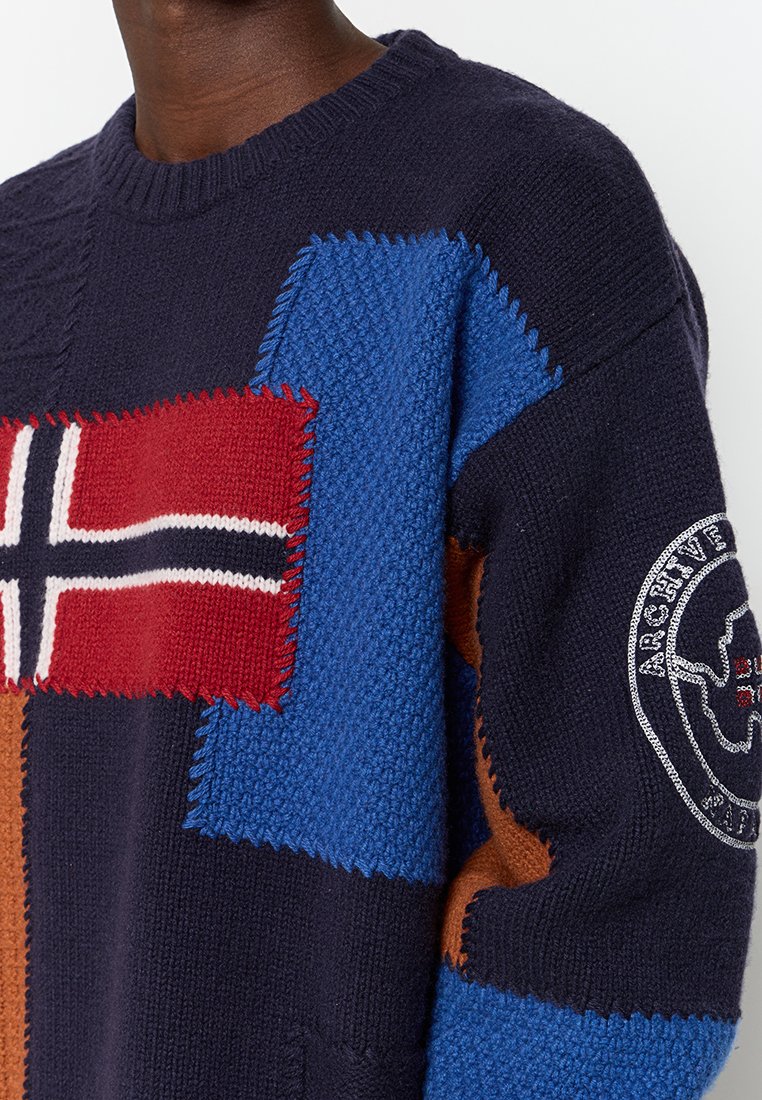 Jersey de punto con color base azul marino, con parches en azul, rojo y marrón. Detalles texturizados y un collar redondo con un parche de logo.