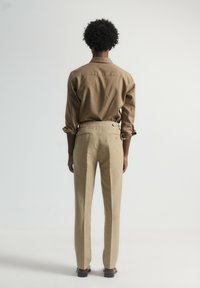 Camisa de algodón marrón con mangas remangadas, combinada con pantalones beige claros que cuentan con dos bolsillos traseros y un corte ajustado.