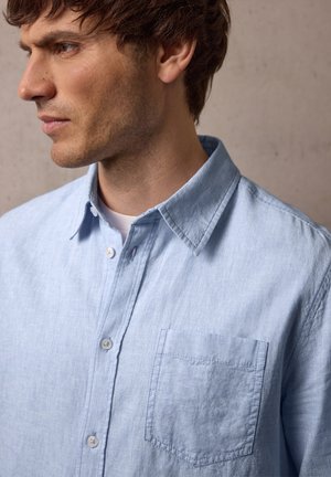 Uomo con camicia azzurro chiaro con taschino sul petto, che guarda a sinistra su sfondo neutro.