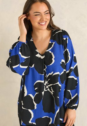 Uśmiechnięta kobieta z długimi brązowymi włosami, ubrana w niebieską bluzkę z dużymi czarno-białymi kwiatowymi wzorami, dotykająca włosów jedną ręką.