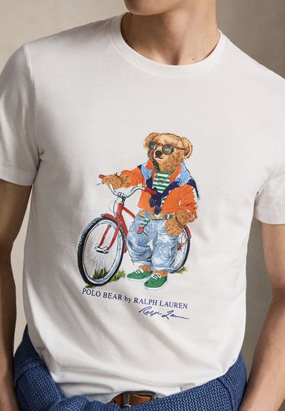 Bjørn med solbriller, oransje jakke, stripete skjorte, jeans og grønne joggesko, stående ved siden av en rød sykkel på en hvit t-skjorte.