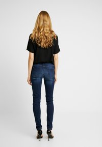Slim-fit tmavě modré džíny spárované s volným černým topem, pohled zezadu. Vlasy jsou vlnité a k délce ramen, obuví jsou černé lodičky.