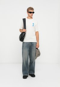 Young man wearing black sunglasses, white t-shirt with blue print, wide-leg jeans, black shoes, holding gray cap and black shoulder bag.