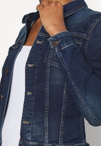 Jeansjacka i mörkblått med en åtsittande design, klassisk krage, knappstängning och slitna detaljer på framsidan och ärmarna.