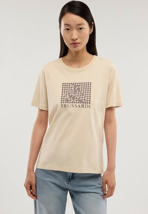 Mujer con cabello largo y oscuro, vestida con una camiseta beige de Trussardi con un logo a cuadros y jeans azul claro, de pie frente a un fondo liso.