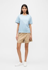 Γυναίκα που στέκεται με το σώμα της στραμμένο προς τα εμπρός, φορώντας ένα ανοιχτό μπλε T-shirt, μπεζ σορτς και λευκά αθλητικά παπούτσια, μπροστά σε ένα απλό ανοιχτό φόντο.