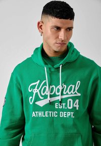 Haut à capuche vert en tissu doux, doté d'une poche kangourou à l'avant, avec le texte imprimé en blanc "Kaporal EST. 04 ATHLETIC DEPT." et des cordons de serrage ajustables.