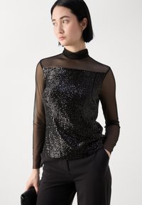 Schwarzes Pailletten-Oberteil mit einem hohen Kragen und transparenten langen Ärmeln, das ein strukturiertes Design aufweist. Kombiniert mit maßgeschneiderten schwarzen Hosen und einer Clutch.