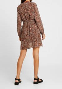 Robe marron à manches longues, longueur genou, avec un imprimé floral blanc et noir. Dispose d'une ceinture à nouer à la taille et d'un tissu léger et transparent.
