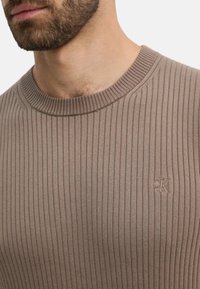 Suéter de punto acanalado en beige con cuello redondo y logotipo en relieve en el pecho. Presenta una textura suave y un diseño ajustado.