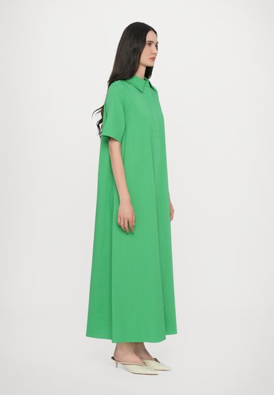 Zielona maxi sukienka z krótkimi rękawami, o luźnym kroju, z jedną kieszenią z przodu. Noszona z białymi, spiczastymi klapkami.