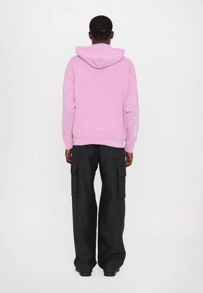 Pink hoodie with a hood, featuring long sleeves and text detail on the left arm, paired with loose black cargo pants and boots.
