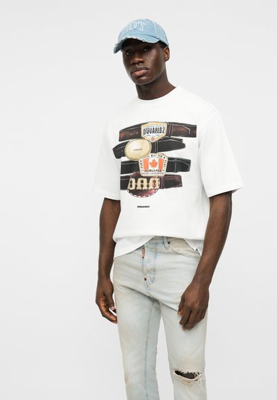 Νέος άνδρας που φοράει λευκό μπλουζάκι με σχέδιο και ανοιχτόχρωμο σκισμένο τζιν, συνδυασμένο με ανοιχτό μπλε τζιν καπέλο, στέκεται μπροστά σε λευκό φόντο.