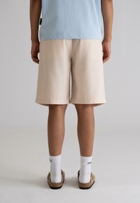 Beige shorts made of smooth fabric, knee-length with a straight cut. Worn with white crew socks and brown slip-on shoes. Minimalist design.