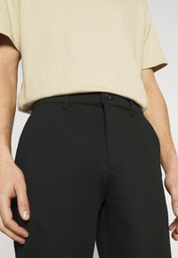 Schwarze Hose aus glattem Stoff, mit schmaler Passform, einfacher Knopfverschluss und Fronttaschen. Kombiniert mit einem hellbeigen T-Shirt.