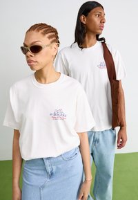 Camisetas de algodón blancas con un estampado gráfico, combinadas con faldas de denim azul claro y jeans. Los accesorios incluyen gafas de sol y un bolso marrón.
