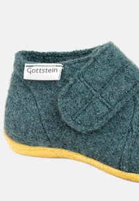 Miękkie, turkusowe wełniane kapcie z żółtą podeszwą oraz etykietą marki Gottstein, z przeszyciami i zapięciem na rzep.