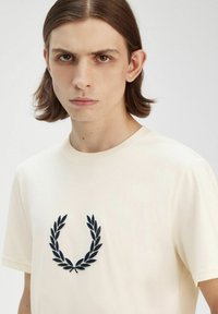 Camiseta de color crema hecha de tela suave con un diseño de laurel bordado en azul oscuro en el pecho, mangas cortas y cuello redondo.