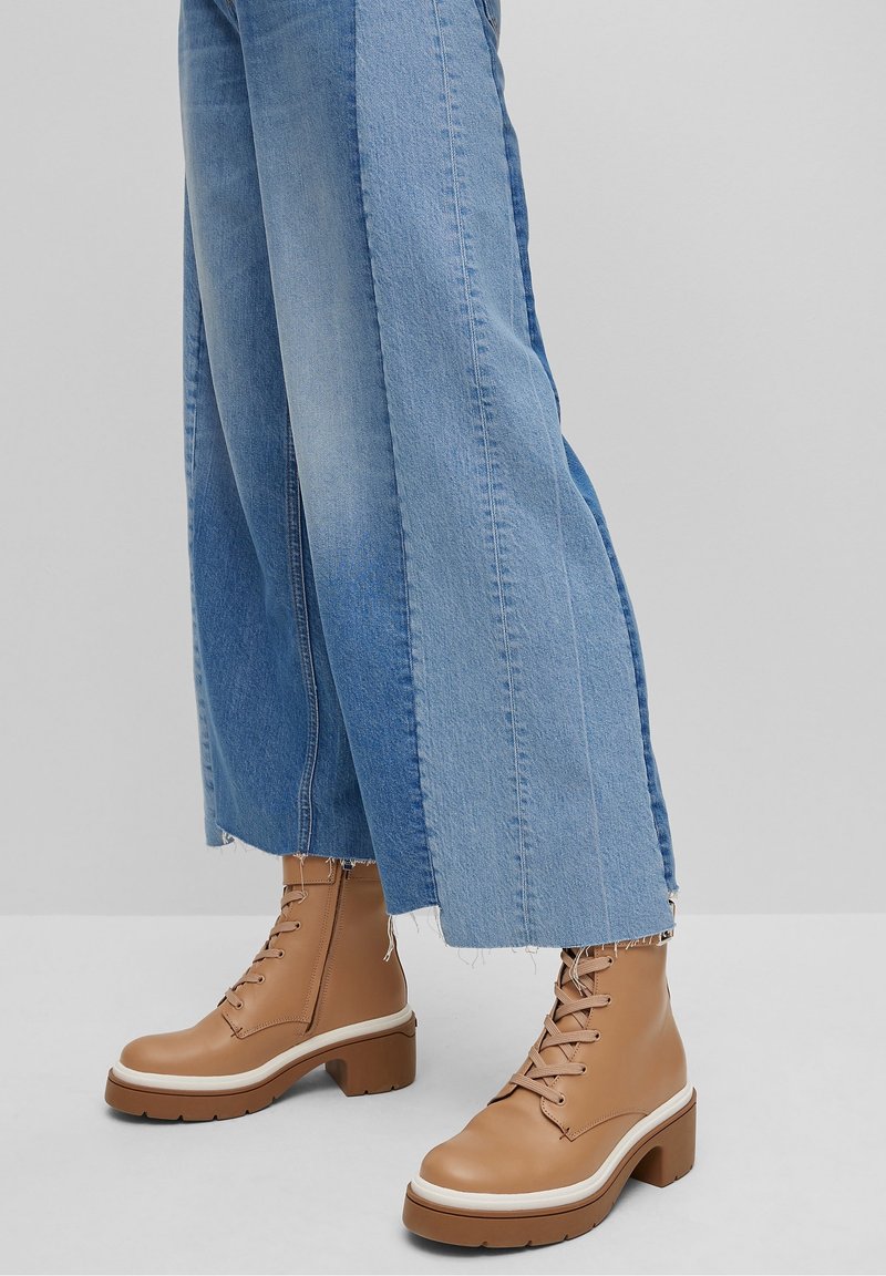 Botas de cuero marrón con cordones, con una suela blanca gruesa y punta redondeada, combinadas con unos vaqueros anchos de color azul claro con un dobladillo sin rematar.