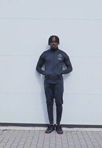 Mustad sportlik track suit, mis on valmistatud siledast kangast, sisaldab lukuga jakki ja kitsenevaid pükse, rinnal on logoga detail. Mustad tenniseketid.