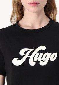Černé bavlněné tričko s velkým bílým stylizovaným logem "Hugo" na přední straně. Krátké rukávy a kulatý výstřih.