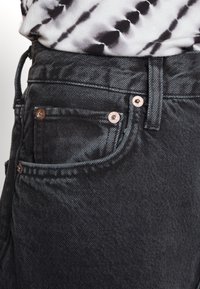 Gros plan sur la poche d'un jean noir en denim avec des rivets métalliques et une partie d'un t-shirt tie-dye blanc et noir rentré à l'intérieur.
