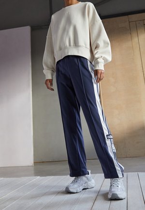 Osoba ubrana w białą, oversize'ową bluzę, granatowe spodnie dresowe z białymi paskami i logo adidas oraz białe masywne sneakersy, stojąca w pomieszczeniu.