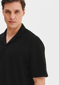 Czarna koszula z krótkim rękawem wykonana z teksturowanej tkaniny, z kołnierzykiem i zapięciem na guziki. Design jest prosty i dopasowany.