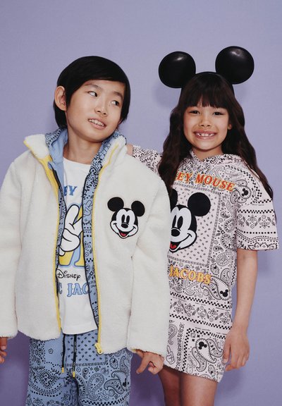 Twee kinderen die glimlachen, waarvan er één een witte fleece jacket met een Mickey Mouse patch draagt, en de ander een Mickey Mouse jurk met zwarte muizenoortjes.
