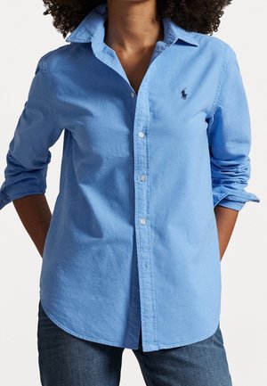 Femme portant une chemise bleu clair boutonnée avec manches retroussées et un jean foncé, debout les mains derrière le dos.