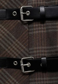 Tissu à carreaux marron avec des sangles en cuir noir. Boucle et détails en métal argenté pour un maintien sûr. Surface texturée avec des lignes.