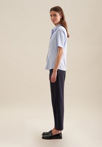 Camicia azzurro chiaro a maniche corte con colletto button-down, abbinata a pantaloni neri su misura e mocassini neri, in piedi davanti a uno sfondo neutro.