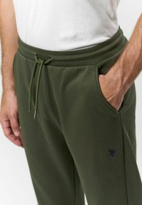 Olivové jogger nohavice s nastaviteľným pásom na šnúrku, bočnými vreckami a decentne vyšitým logom na ľavej nohe. Mäkká zmes bavlny.