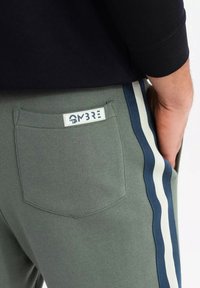 Graue Sweatpants mit seitlichen Streifen in Marineblau und Weiß, mit einer Gesäßtasche, die ein Etikett mit der Aufschrift "OMBRE" trägt, und einer glatten Textur.