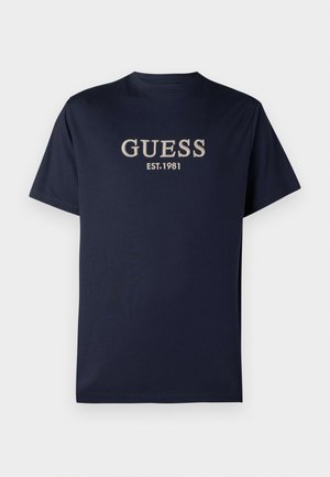 T-shirt azul marinho de manga curta com "GUESS EST. 1981" bordado em branco no peito, exibido sobre um fundo simples.
