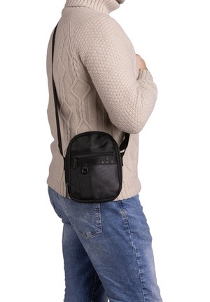 Borsa a tracolla nera con un design strutturato, diverse tasche con zip e superficie testurizzata. Cinghia regolabile attaccata, indossata di lato.