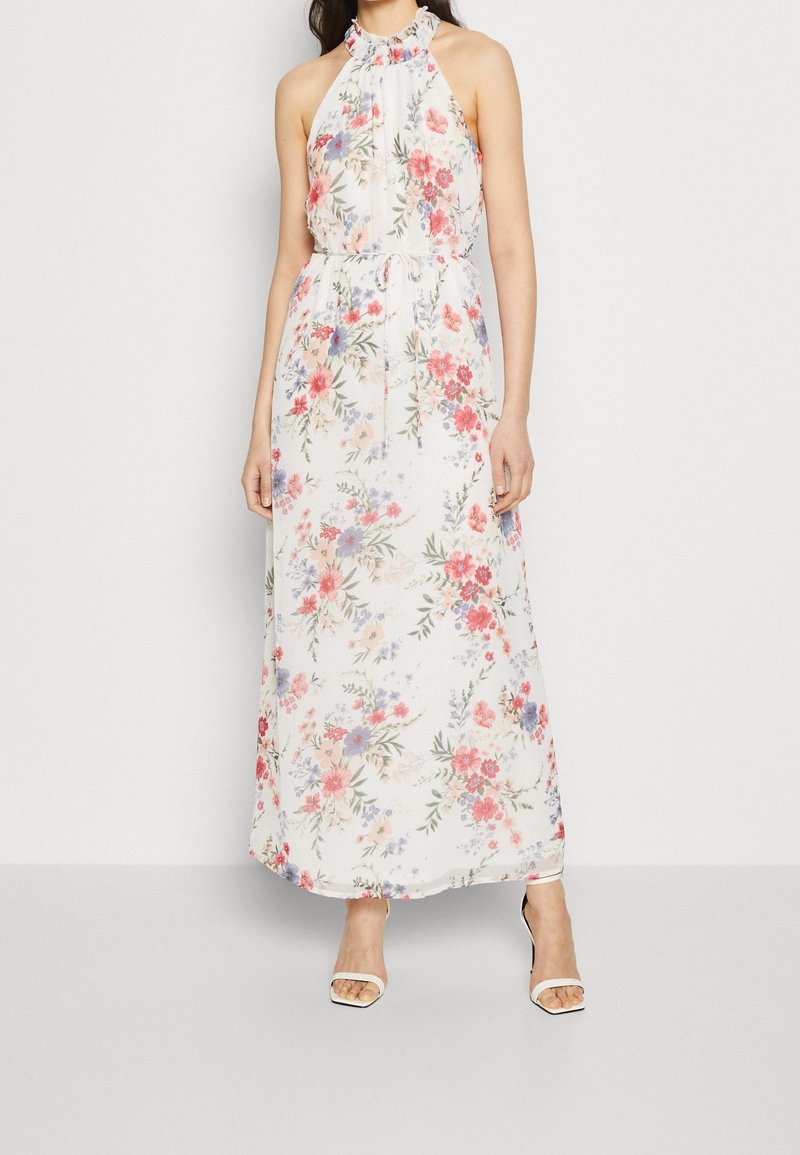 Femme portant une robe maxi fleurie blanche sans manches avec un col halter et des talons ouverts blancs sur un fond uni.