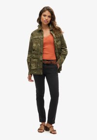 Chaqueta de camuflaje verde oliva con múltiples bolsillos, camiseta sin mangas naranja, pantalones negros y sandalias de tacón marrones con acentos decorativos.