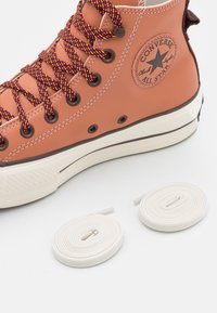 Oranžové kožené vysoké tenisky s fialovými a žlutými vzorovanými tkaničkami. Součástí jsou také dvě bílé ploché tkaničky vedle boty. Klasická gumová podrážka.