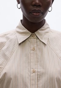 Cămașă bej cu dungi, cu guler clasic, nasturi în față și mâneci raglan. Textură netedă, cu doi nasturi vizibili la decolteu.