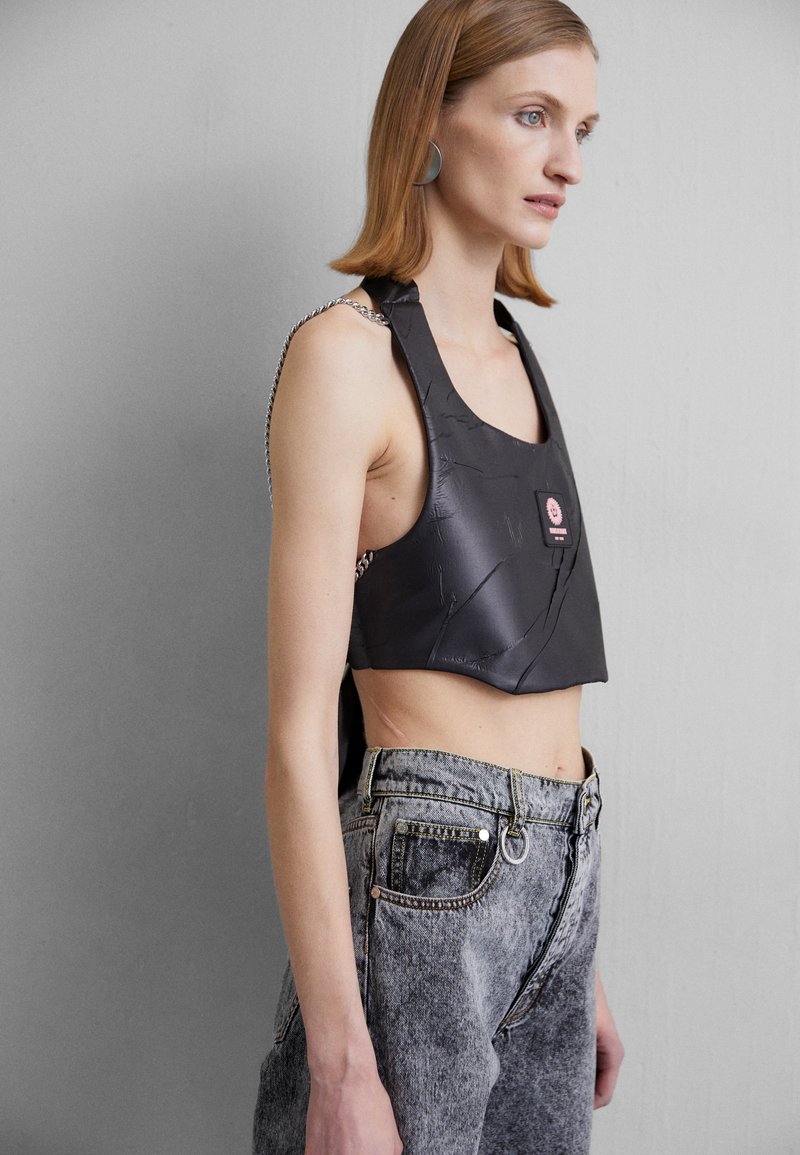 Black cropped top made of textured material with a square neckline, chain accents on straps, and a prominent pink label. Paired with gray jeans.