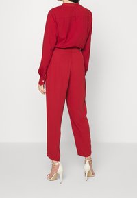 Mono rojo con mangas largas, un corte relajado y una cintura alta. Presenta una cremallera en la parte trasera y botones en los puños. Combinado con tacones blancos.