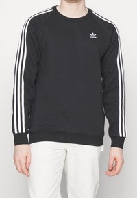 En person som bär en svart Adidas sweatshirt med vita ränder på ärmarna och logotypen på bröstet, i kombination med vita byxor.