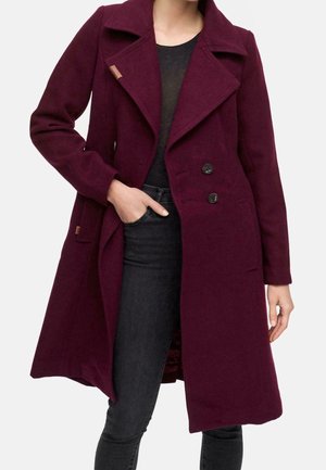 Frau trägt einen dunkelvioletten, zweireihigen Wollmantel über einem schwarzen Top und dunklen Jeans, mit einer Hand in der Manteltasche.