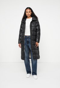 Sort pufferjakke med et langt design, skinnende materiale, knaplukning i kraven og lodrette syninger, parret med løse blå jeans og hvide sneakers.