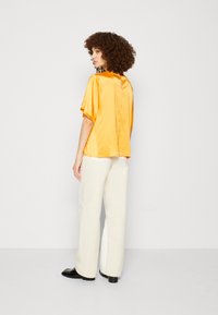 Blusa gialla in satin a maniche corte con un'apertura a chiave sul retro, abbinata a pantaloni bianchi a gamba larga con vita alta e scarpe nere. Capelli ricci visibili.