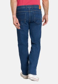 Jean en denim bleu pour homme avec une coupe droite, cinq poches et un patch en cuir marron sur la taille arrière. Porté avec des baskets blanches.