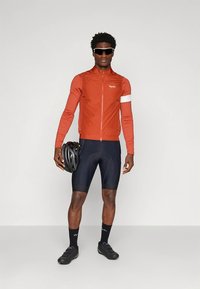 Rode lichtgewicht fietsjas met een witte accent, zwarte gevoerde korte broek, zwarte schoenen en een zwarte helm, gedragen door een staande fietser.