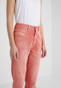 Personne portant un jean rose corail délavé taille haute et un top blanc sans manche uni, debout devant un fond gris clair.